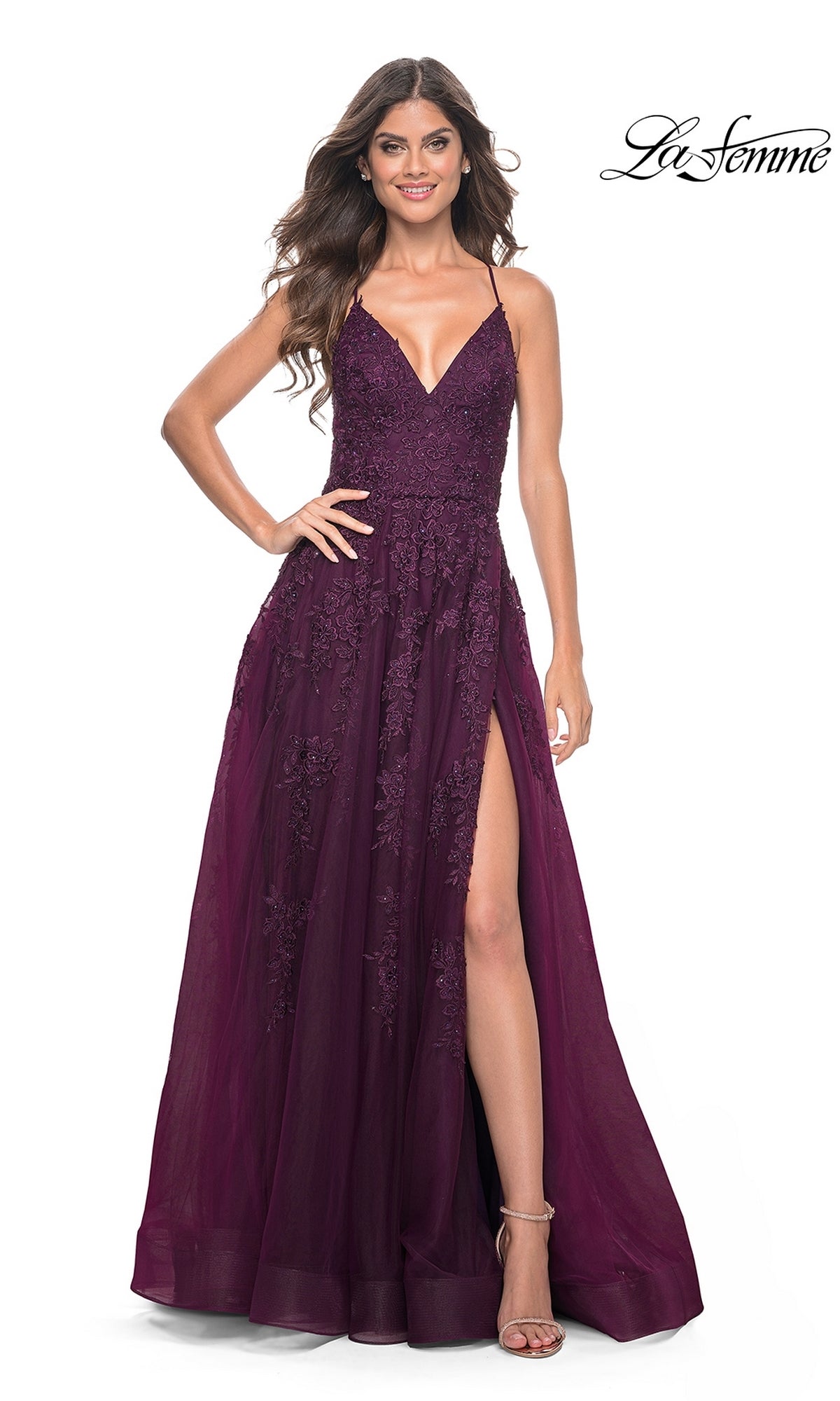A woman poses with one hand on her hip in the La Femme Long Lace A-Line Prom Dress 32303, featuring a sleeveless design, deep purple hue, lace details, V-neckline, and high slit against a white background.